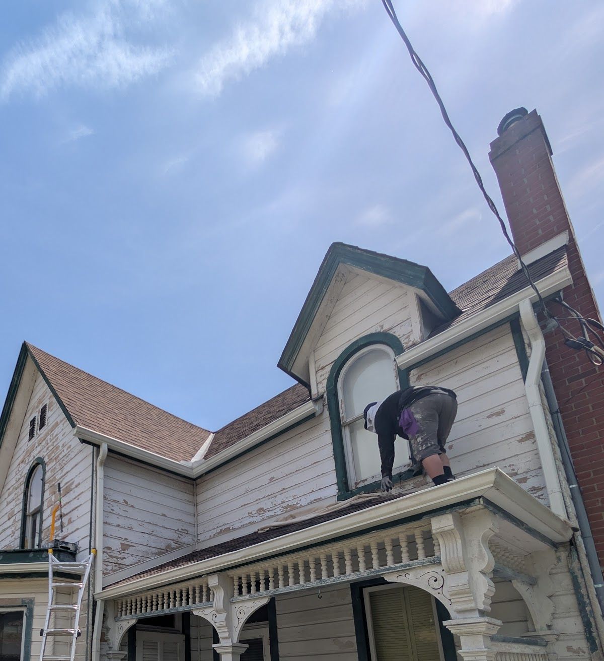 Painter on the porch roof of a century home with peeling siding, dark green trim, and a brick chimney, while the exterior repaint is still in progress